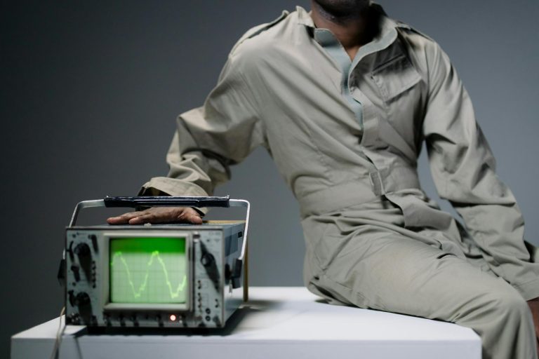 A person in coveralls sits beside an oscilloscope displaying a waveform in a studio setting.